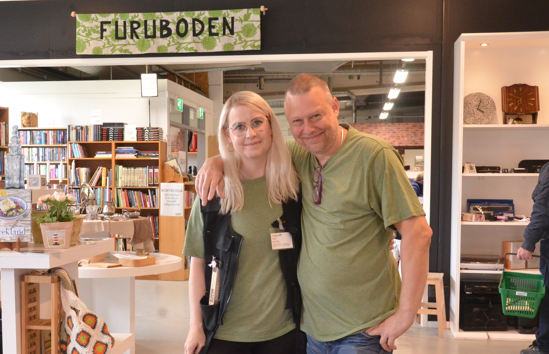 En kvinna och en man står i en second hand butik och tittar in i kameran